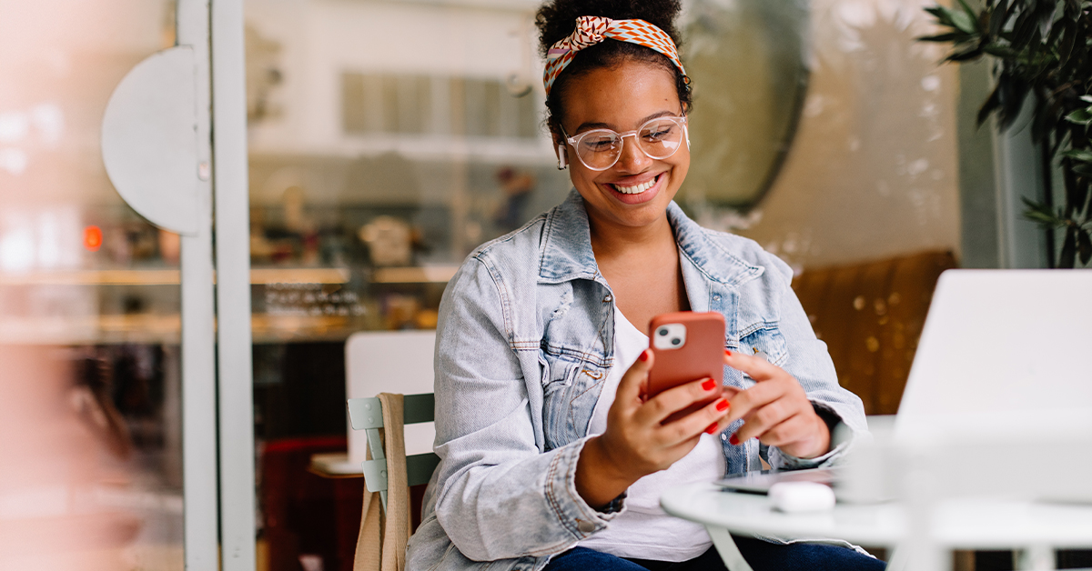 Smiling woman sitting at a café table using her smartphone, with a laptop and coffee nearby